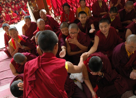 Young Tibetan Buddhist nun handing out initiation nectar for nuns to drink, traditional red blindfolds, maroon robes, malas, front porch, monks in background, Sakya Lamdre, Tharlam Monastery of Tibetan Buddhism, Boudha, Kathmandu, Nepal