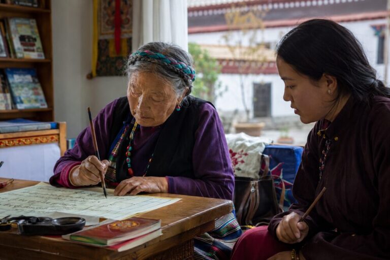 An older Tibetan woman practices calligraphy with a brush while a younger woman studies beside her.